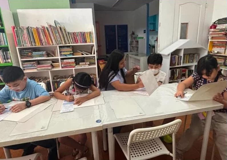 Five children around a table in the school library, each working on a large sheet of their own